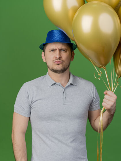 穿严格的年轻人戴着派对帽拿着气球孤立在绿色严格绿色抱着