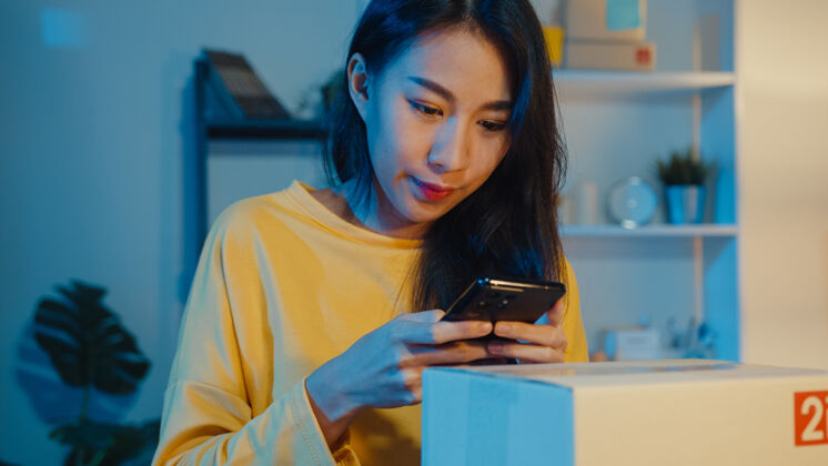 办公室年轻的亚洲妇女使用智能手机在包裹上拍摄条形码照片 以便将货物运送给客户手机工作到很晚晚上