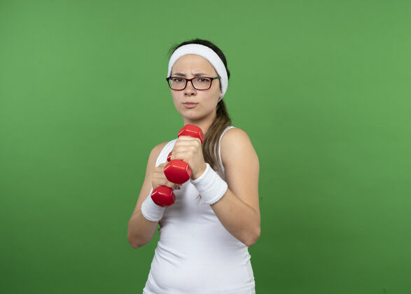 穿戴着眼镜 戴着头带和腕带的严肃的年轻运动女性在绿色的墙上举着孤立的哑铃女孩人光学