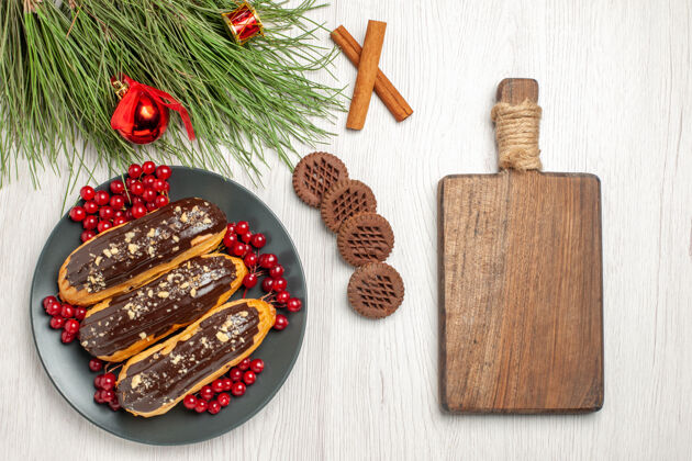 肉桂俯瞰图：灰色盘子上的巧克力和葡萄干饼干交叉着桂皮和松叶 白色木桌上放着圣诞玩具和砧板十字架木板桌子