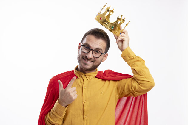 王冠微笑的超级英雄戴着眼镜 披着红色斗篷 头上戴着王冠 指着隔离在白墙上的前方人眼镜举行