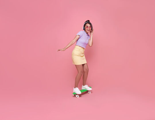 乐趣年轻的亚洲少女戴着无线耳机在粉红色的墙上玩滑板 听音乐时尚女人肖像