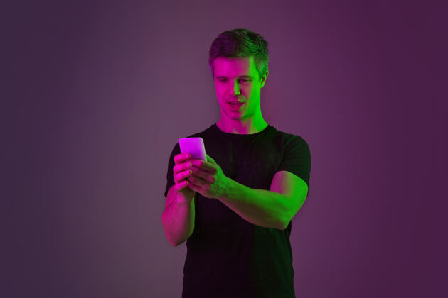 人使用智能手机 自拍 vlog霓虹灯下紫色工作室背景上的白种人肖像穿着黑色衬衫的漂亮男模人类情感的概念 面部表情 销售 广告手主管公司