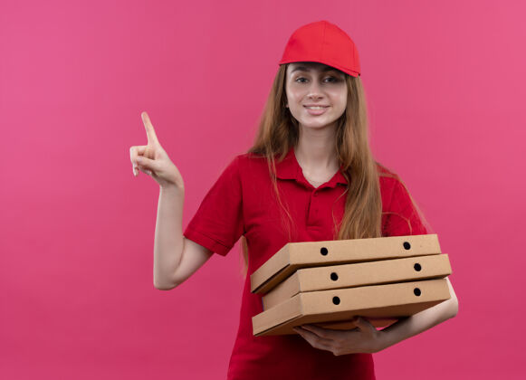 年轻微笑着的年轻送货女孩穿着红色制服拿着包裹 指着孤立的粉红色空间和复印空间女孩盒子空格