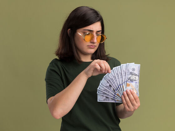 绿无知的年轻漂亮的高加索女孩 戴着太阳眼镜 拿着橄榄绿的钞票看着眼镜无知年轻