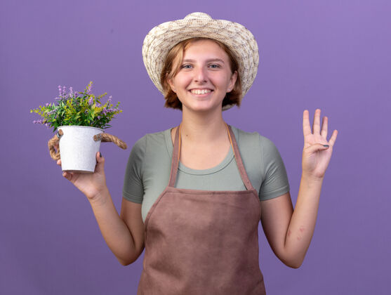 四微笑着的年轻的斯拉夫女园丁戴着园艺帽 手里拿着花盆里的花 四个手指在紫色上做手势花四四