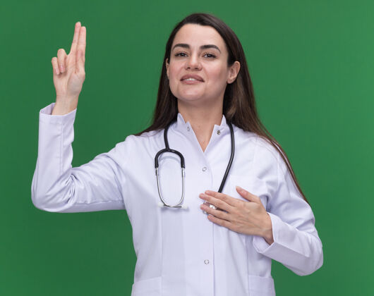 年轻高兴的年轻女医生穿着医用长袍 用听诊器在绿色上做宣誓手势女医生手势