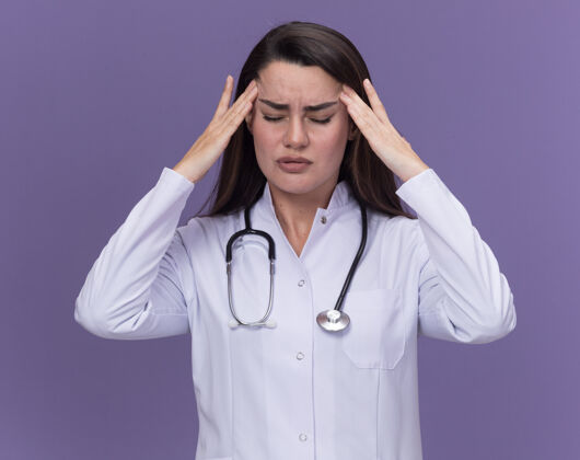疼痛疼痛的年轻女医生穿着带听诊器的医用长袍 双手放在头上 闭着眼睛站在紫色上年轻女性医生