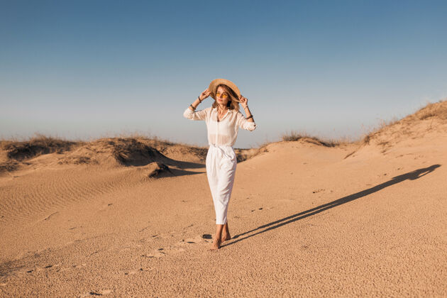 美丽一个穿着白色衣服 戴着草帽 在沙漠沙滩上行走的时尚美女旅游模特沙漠