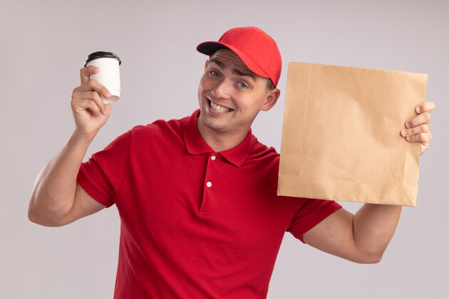 人带着微笑的年轻送货员 穿着制服 戴着帽子 手里拿着一个纸制食品包 白墙上隔着一杯咖啡人年轻人人