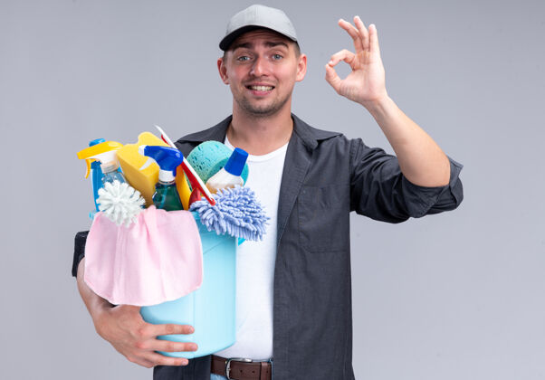 帽子面带微笑的年轻帅气的清洁工 穿着t恤 戴着帽子 手里拿着一桶清洁工具 在白色的墙上显示出良好的姿势人工具公民