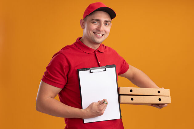 感觉微笑着的年轻送货员穿着制服 戴着帽子 拿着披萨盒 剪贴板被隔离在橙色的墙上递送盒子帽子