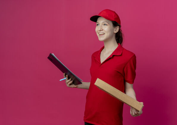 深红色快乐的年轻漂亮的送货女孩 穿着红色制服 戴着帽子站在侧视图中 手里拿着披萨包和剪贴板 手里拿着一支笔 笔直地孤立在深红色的背景上 还有复印空间直钢笔查看
