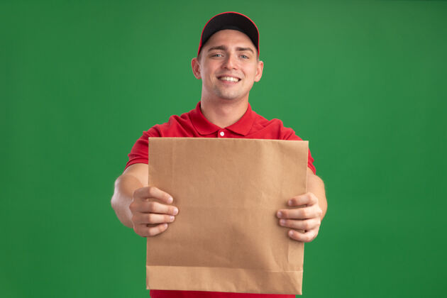 穿着面带微笑的年轻送货员身穿制服 头戴鸭舌帽 手拿纸质食品包 站在绿色的墙前表情公民制服