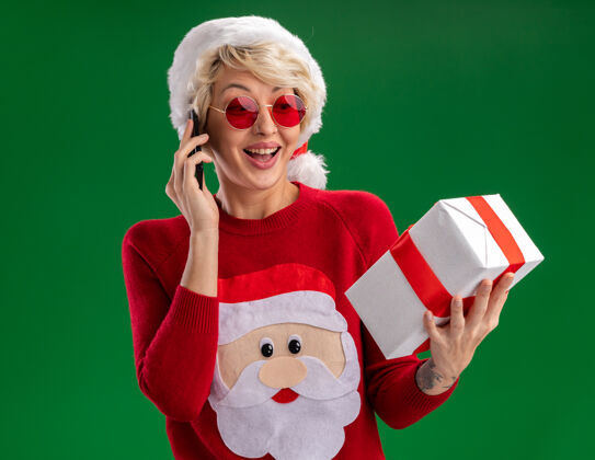 金发快乐的金发女郎戴着圣诞帽 穿着圣诞老人的圣诞毛衣 戴着眼镜 拿着礼物包 在绿色的背景下 隔着电话聊天欢乐包装礼物