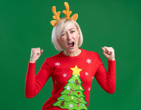 绿色快乐的中年金发女人戴着圣诞驯鹿鹿角头带和圣诞毛衣看着相机眨眼做是的手势隔离在绿色背景上手势金发中年