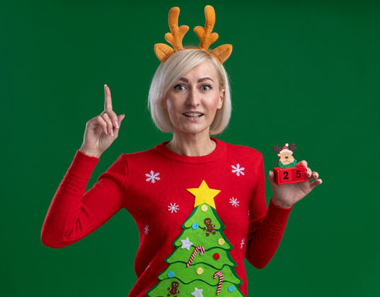 圣诞快乐快乐的中年金发女人戴着圣诞驯鹿鹿角头带和圣诞毛衣拿着圣诞驯鹿玩具和日期看着相机指向上孤立的绿色背景玩具日期毛衣