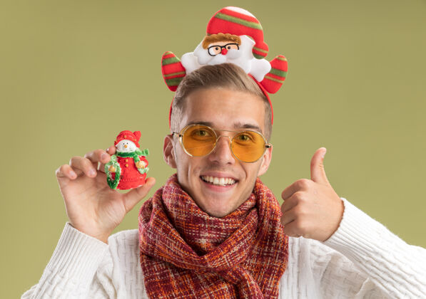 拇指快乐的年轻帅哥戴着圣诞老人的头带和围巾看着拿着雪人圣诞饰品的相机 在橄榄绿的背景上孤立地竖起大拇指显示头带圣诞老人
