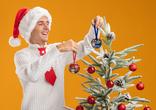 圣诞快乐快乐的年轻帅哥戴着圣诞帽 打着圣诞老人的领带站在圣诞树旁 用橙色背景上孤立的圣诞球装饰圣诞树圣诞老人站圣诞树