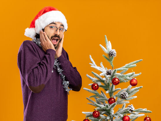 脸颊兴奋的年轻帅哥站在圣诞树旁 戴着圣诞帽 脖子上戴着花环 手放在脸颊上 橘黄色的背景隔离开来小伙子手橘子