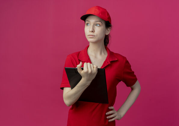 背景年轻漂亮的送货女孩 穿着红色制服 戴着帽子 手里拿着剪贴板 手放在腰上 一边看着一边 在深红色的背景上孤立着 还有复印空间帽子举行女孩