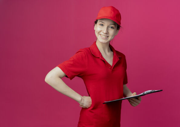 帽子微笑着的年轻漂亮的送货女孩 穿着红色制服 戴着帽子 手放在腰上 在深红色背景上 留有复印空间剪贴板腰深红色