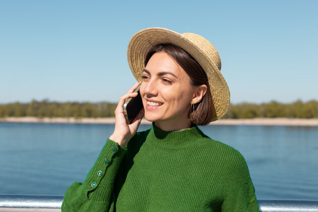 放松穿着绿色休闲毛衣 戴着帽子的时髦女人在户外的桥上 在温暖阳光下的夏日用手机聊天手机蓝色电话