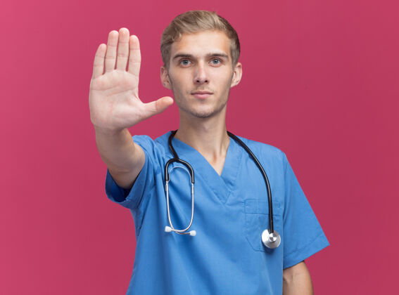 表演严格的年轻男性医生穿着医生制服 听诊器显示停止手势隔离在粉红色的墙上制服站住男性