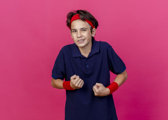 脸自信的年轻帅气的运动男孩戴着头带和护腕 戴着牙套 握紧拳头 看着前面的粉红色墙壁上的复制空间隔离运动握紧人