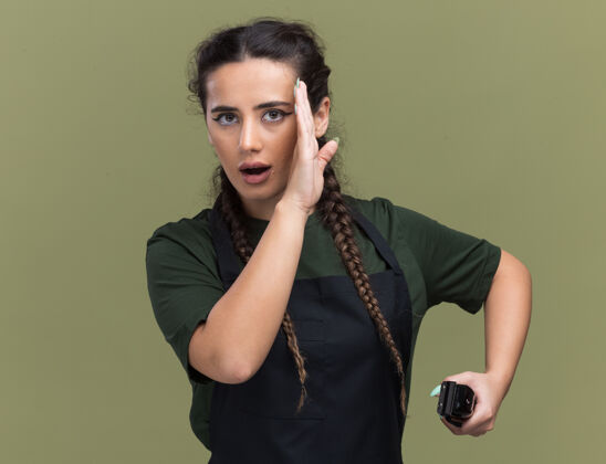 女可疑的年轻女理发师穿着制服拿着理发器在橄榄绿的墙上窃窃私语制服年轻窃窃私语