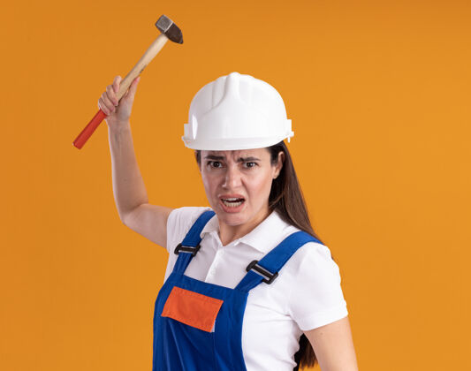 建筑工人愤怒的穿着制服的年轻建筑妇女手持铁锤孤立在橙色的墙上女人年轻拿着