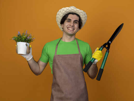 持有年轻的男园丁面带微笑 穿着制服 戴着园艺帽 戴着手套 拿着剪子 把花盆里的花隔离在橘黄色上穿年轻制服