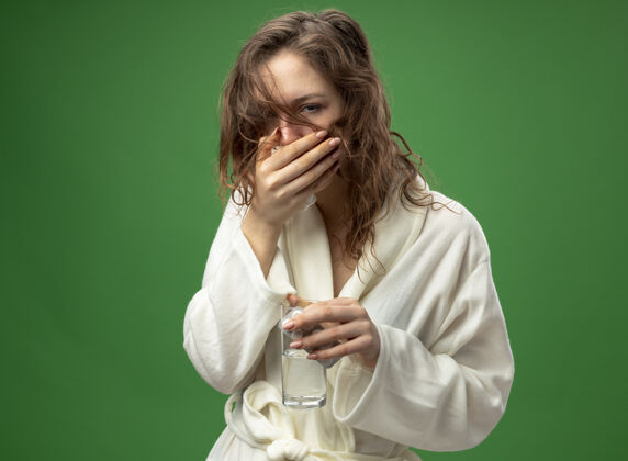 病咳嗽的年轻生病的女孩穿着白色长袍 拿着一杯水和药片 用手捂着嘴隔离在绿色上幼绿丸