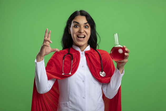 绿色兴奋的年轻超级英雄女孩穿着医用长袍 手持装有听诊器的化学玻璃瓶 里面装满了红色液体 绿色上显示出孤立的大小显示化学红色