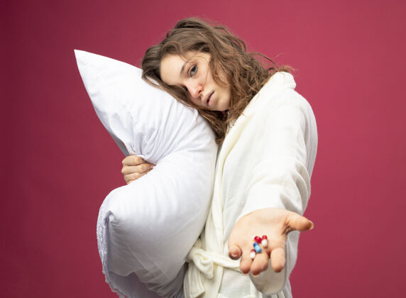 粉色体弱多病的年轻女孩穿着白色长袍抱着抱枕拿着隔离在粉色上的药丸女人人女性