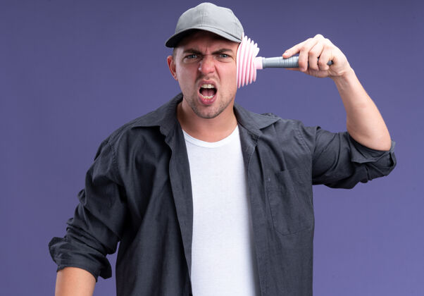 帽子愤怒的年轻帅气的清洁工穿着t恤 戴着帽子 头上拿着柱塞 隔离在紫色的墙上愤怒持有年轻