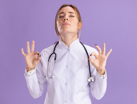 医学年轻的女医生闭着眼睛 穿着带听诊器的医用长袍 在紫色的墙上显示冥想的姿势穿手势封闭