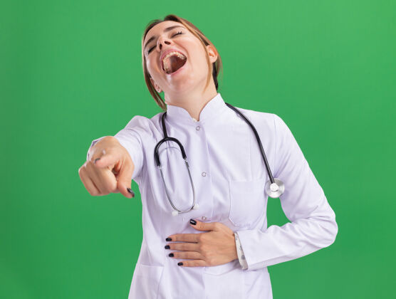 大笑年轻的女医生穿着医用长袍 带着听诊器 笑嘻嘻地抓住斯托姆奇 在绿色的墙上给你们展示隔离的姿势长袍手势医生