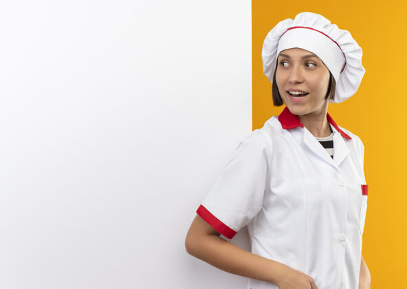精益快乐的年轻女厨师身着厨师制服 倚在白色的墙上 看着孤立在橙色的一面欢乐墙壁制服