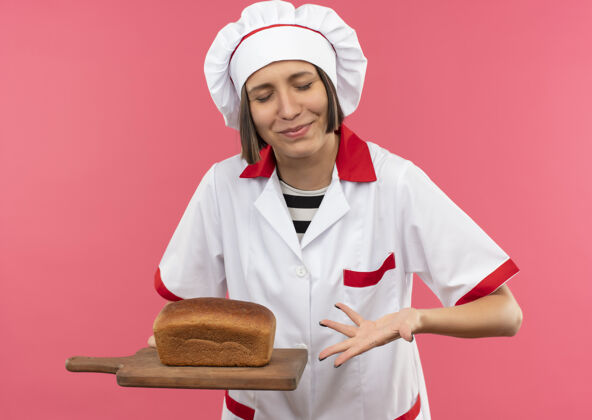 眼睛身着厨师制服的年轻女厨师手捧着面包 用手指着砧板 闭着眼睛 孤立地站在粉色背景上烹饪封闭拿着