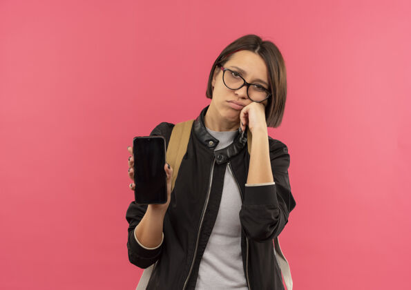 眼镜未出租的年轻女学生戴着眼镜 背着包拿着手机 手放在脸颊上 隔离在粉色背景上 留有复印空间年轻手机背