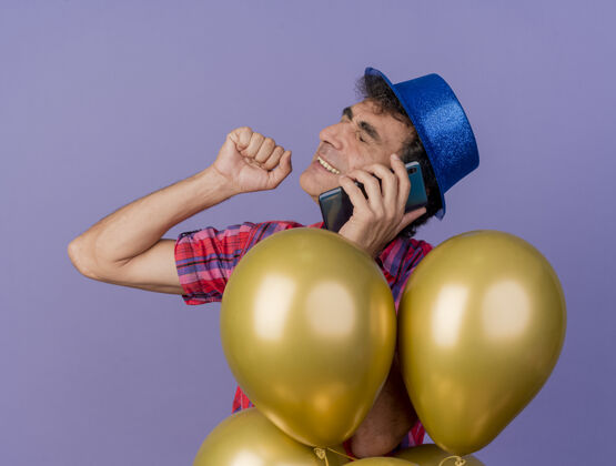 衣服微笑着的中年党人戴着党的帽子站在气球后面讲电话保持拳头在空中闭着眼睛孤立在紫色的墙上气球人眼睛