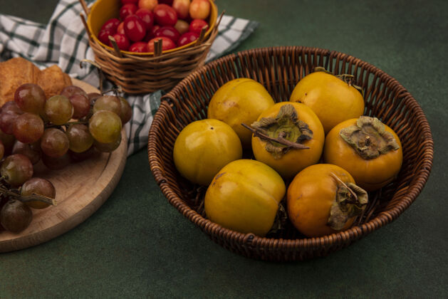 景观柿子水果的顶视图在一个桶上 葡萄放在绿色背景的格子布上的木制厨房板上木头新鲜葡萄