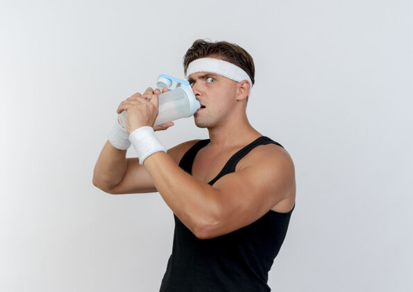 帅气印象深刻的年轻英俊的运动男子戴着头带和腕带饮用水从水瓶上孤立的白色年轻腕带喝酒