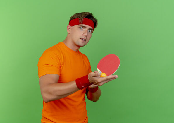 帅气印象深刻的年轻英俊的运动男子戴着头带和手环举行乒乓球拍和球隔离在绿色穿持有球拍