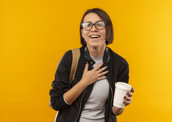 眼镜快乐的年轻女学生戴着眼镜 背着袋子 手里拿着塑料咖啡杯 手放在胸前 隔离在橘色上学生包抱着