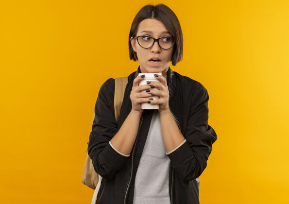 包印象深刻的年轻女学生戴着眼镜 背着塑料袋拿着咖啡杯 看着边上孤立的橙色塑料背部橘子
