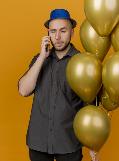 抱着年轻帅气的斯拉夫派对小伙子戴着派对帽拿着气球在电话里聊天看着橙色背景下的孤零零小伙子年轻人气球