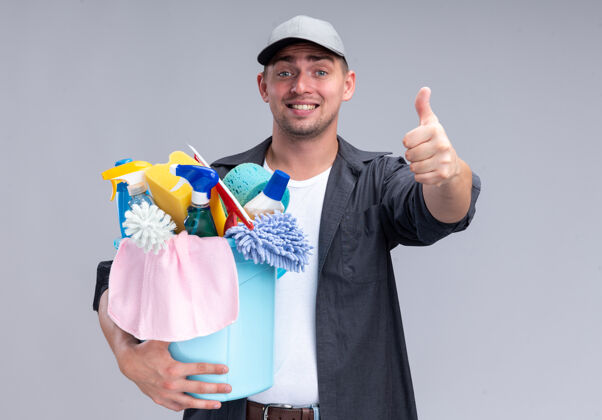 帽子微笑着的年轻帅气的清洁工 穿着t恤 戴着帽子 手里拿着一桶清洁工具 在白色的墙上孤立地竖起大拇指工具微笑显示