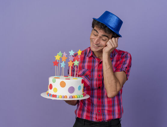 紫色微笑的中年白种人党男子戴着党的帽子举行和看生日蛋糕把手放在脸上孤立的紫色背景与复制空间手蛋糕复制